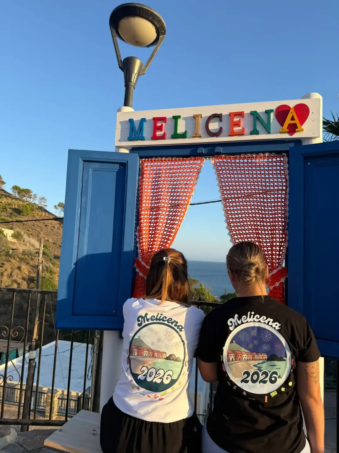 Chicas con las camisetas oficiales de Melicena 2026, frontal. Tenemos la versión en blanco y negro, ambas ponen 'Melicena 2026'.