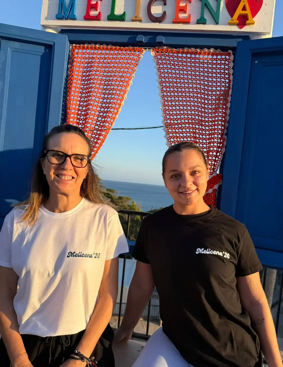 Chicas con las camisetas oficiales de Melicena 2026, frontal. Tenemos la versión en blanco y negro, ambas ponen 'Melicena 2026'.